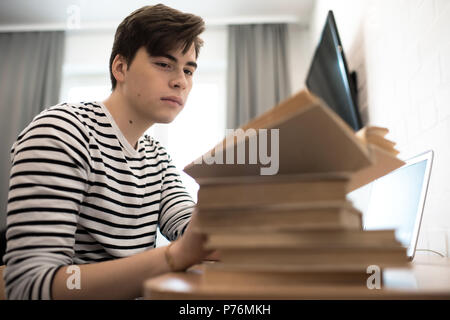 Thoughtful young male sitting at home with pile of books and reading. Stock Photo