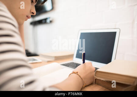 Attractive young male sitting at home at desk with laptop and making notes while reading book. Stock Photo