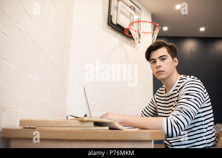 Attractive young male sitting at home at desk with books using laptop computer and looking at camera. Stock Photo