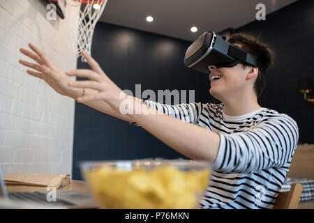 Excited young male extending hands forward while using virtual reality headset sitting at home. Stock Photo