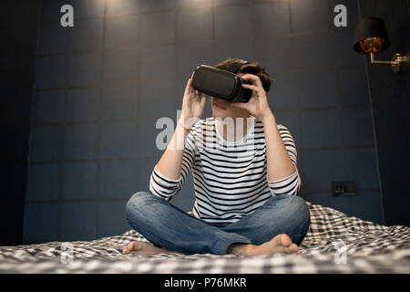 Young male using virtual reality headset while sitting on bed in dark room. Stock Photo