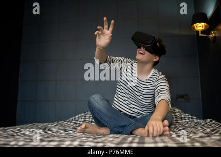 Excited young man extending hand forward while using virtual reality headset sitting on bed in dark room. Stock Photo