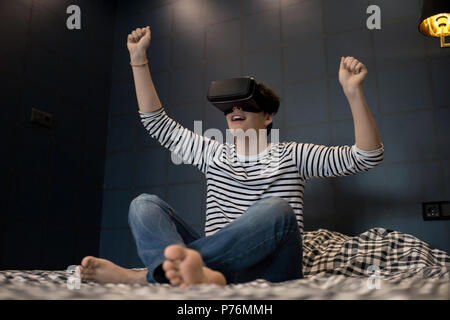 Young male raising arms while using virtual reality headset sitting on bed in dark room. Stock Photo