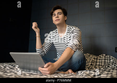 Attractive young man sitting on bed with laptop computer and looking away dreamy. Stock Photo