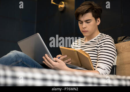 Attractive young male concentrated on reading book while sitting on bed and holding laptop computer. Stock Photo