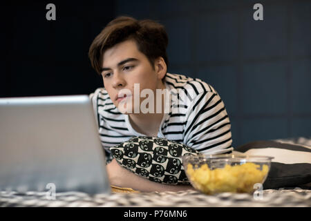 Handsome young male lying on bed with bowl of chips and reading on laptop screen. Stock Photo