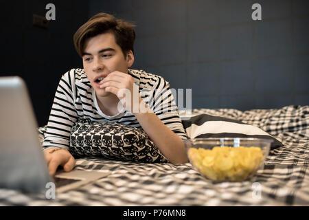Attractive young male lying on bed and eating chips concentrated on using laptop computer. Stock Photo