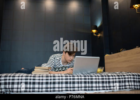 Attractive young male lying on bed with stack of books using laptop computer and taking notes. Stock Photo