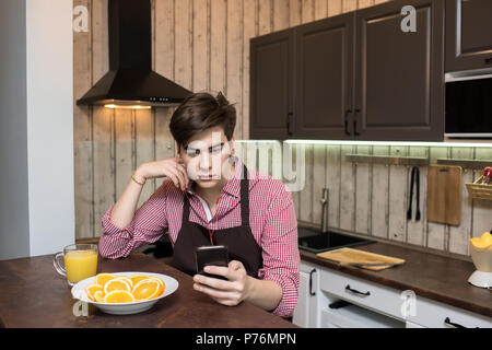 Young man enjoying good music and using smartphone while sitting in cozy kitchen alone. Stock Photo