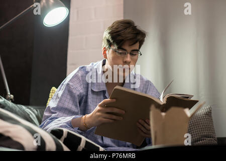 Handsome young man in pajamas and glasses sitting on comfortable sofa and reading nice book. Stock Photo