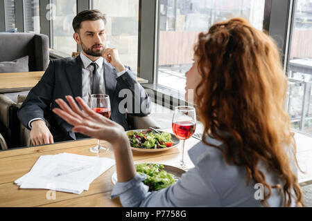 Man and woman having business lunch at restaurant sitting at table eating fresh salad drinking red wine talking discussing project relaxed Stock Photo
