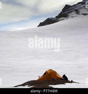 Hiker sitting near orange camping tent in evening snowy mountains on overnight stay. Turkey, Central Taurus Mountains, Aladaglar (Anti-Taurus), platea Stock Photo