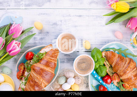Family breakfast of two croissants with arugula and cheese and aromatic coffee, eggs of different colors. A bouquet of tulips in a blue vase. Free space for text. White wooden background Stock Photo