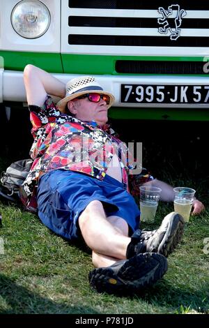 Man in straw hat wearing floral shirt and sunglasses lying down drinking beer and leaning against a van at Isle of Wight festival Stock Photo