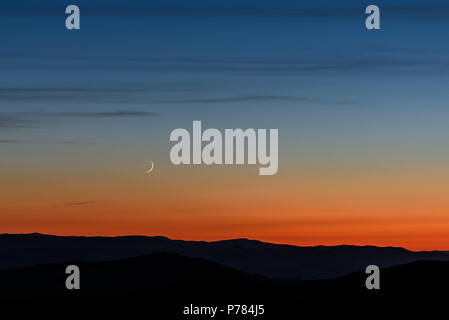 Beautiful evening landscape with a narrow crescent moon and clouds in the dark blue sky above the mountains against the background of the orange sunse Stock Photo