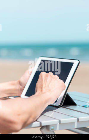 closeup of a young caucasian man on the beach, using a digital tablet, sitting at an outdoors table, with the ocean in the background Stock Photo