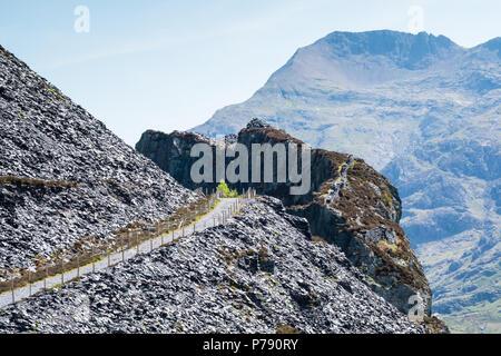 Path to Nant Peris through Dinorwic slate quarry in Llanberis, Wales, with Snowdonia mountain range in background and a hazy blue sky Stock Photo