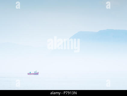 Lone fisherman and red fishing boat on a calm misty day in St George's Channel near Beaumaris on the Isle of Anglesey in Wales with mountain backdrop Stock Photo