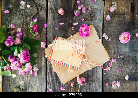 Top view pile of old letters in craft paper envelopes, blank greeting card and wilting tea rose flowers on the old wooden rustic background. Vintage c Stock Photo