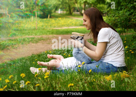 Young pregnant girl sits on lawn in city park. Mother plays with her son. Little boy is lying on lawn. Blur effect Stock Photo