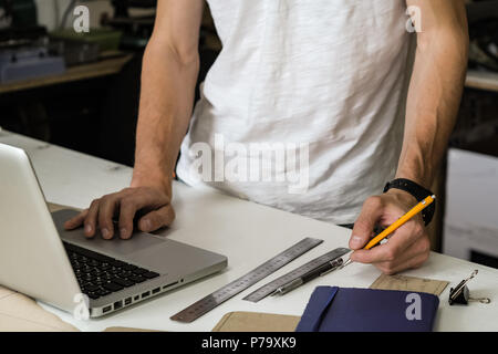 Consumer goods designer at work in workshop. Hands of young male person with pencil and laptop computer at a working table of small manufacture facili Stock Photo