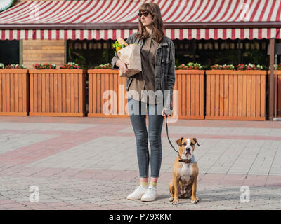 Young pretty woman with dog holds paper bag of groceries in front of market place or vegetable store. Going to shop for food with trained pitbull terr Stock Photo
