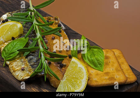 Two pieces of smoked mackerel sprinkled with colorful pepper, garnished with lemon, a fresh sprig of rosemary and basil leaves, on an old wooden board Stock Photo