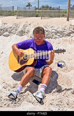 A senior adult male sits on a sandy beach playing a guitar Stock Photo