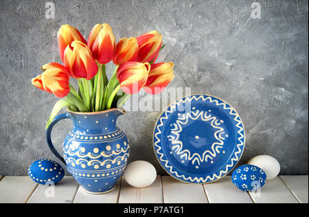 Red-yellow tulips in blue ceramic pitcher with Easter eggs on gray stone background Stock Photo