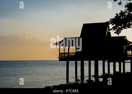 A picturesque scene of the black silhouette of a traditional Malay house on stilts with a tropically-suited roof at the sea during sunset in Malaysia. Stock Photo