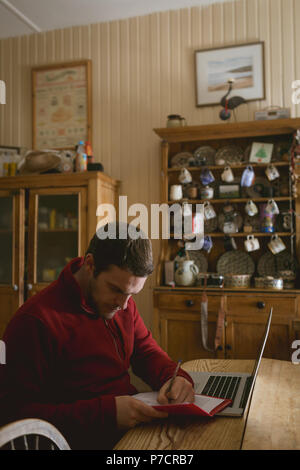 Man writing on a book while using laptop Stock Photo