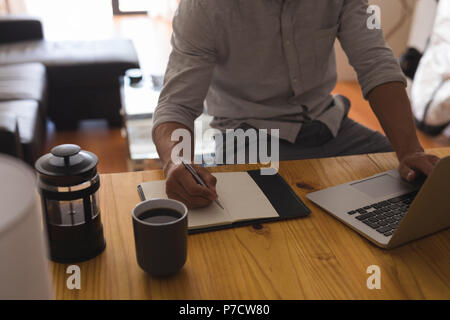 Man writing on a diary while using laptop at home Stock Photo