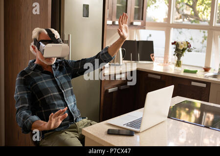Senior man using virtual reality headset with laptop in kitchen Stock Photo