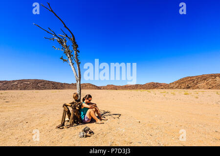 Young woman sitting under a dried tree next to a cairn Lonely Man, Hartmann Valley, Kunene region, Namibia Stock Photo