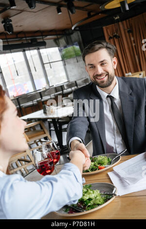 Man and woman having business lunch at restaurant sitting at table eating vegetable salad drinking red wine shaking hands smiling joyful agreement Stock Photo