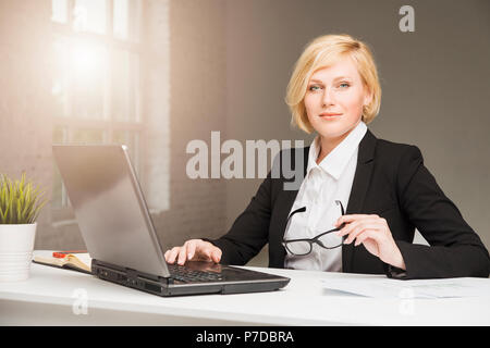 Beautiful blonde adult businesswoman dressed in black stylish suit with eyeglasses working in white wide office on table with laptop Stock Photo