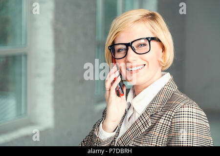 Beautiful blonde adult businesswoman dressed in black stylish suit with eyeglasses talking via smartphone Stock Photo