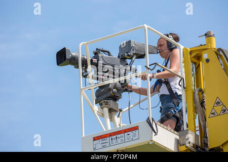 Close up of television camerawoman isolated on elevated platform raised high in midair filming sport event for live broadcast. Blue sky background. Stock Photo