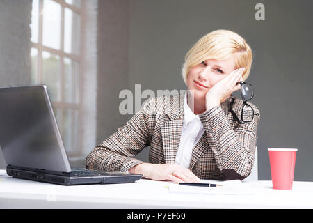 Tired blonde businesswoman dressed in stylish suit sitting at the workplace with laptop computer Stock Photo