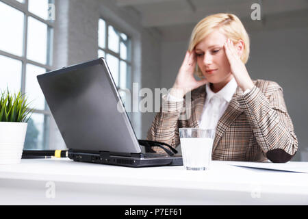 Blonde businesswoman dressed in stylish suit have working headache, using effervescent pill on water in glass, workplace with laptop in white office Stock Photo