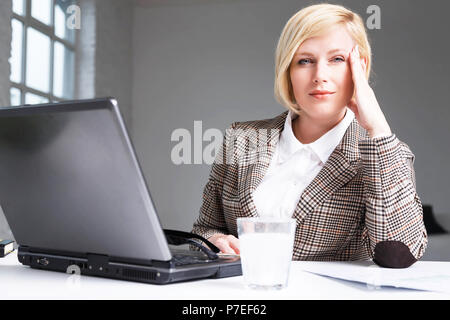 Blonde businesswoman stylish suit have working headache, using effervescent pill on water in glass, workplace with laptop in white office Stock Photo