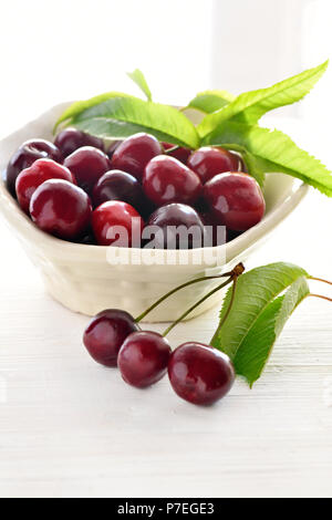 fresh summer cherries in a ceramic dish with greenery on a white colored background, vertical with copy space and room for text overlay Stock Photo