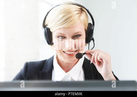 Smiling call center operator, blonde woman dressed in black jacket and white shirt working on laptop via headset in white office Stock Photo