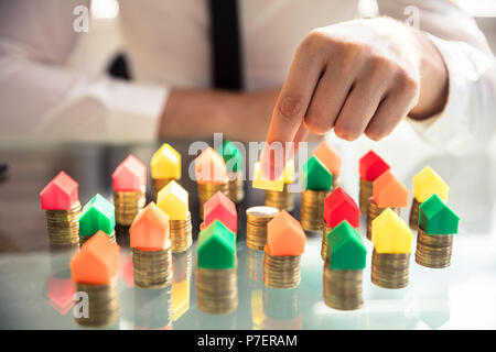 Close-up Of A Person's Hand Placing Multi Colored House Model On Stacked Coins Over Desk Stock Photo