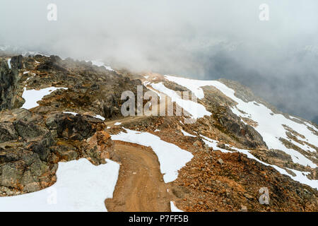 A view from above on a country road, stony road, winding in the mountains between rocks, snowdrifts on rocks, dense fogs in the distance Stock Photo