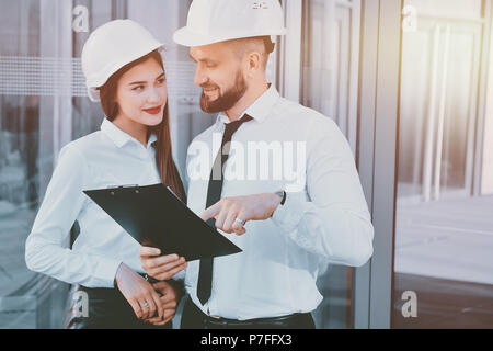 A girl with a man in construction white helmets and white shirts with drawings in hands against a glass office building. Engineers. Planning of constr Stock Photo