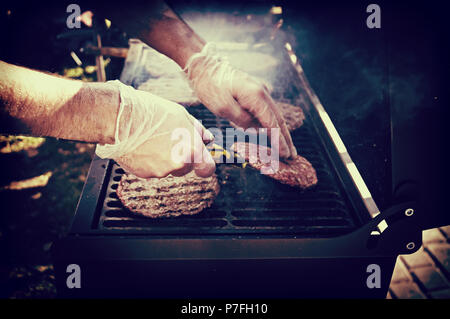 Burgers grilled on charcoal BBQ grill. The chef flips patties roasted on an open fire with a metal spatula. Ingredients for burgers from beef mince Stock Photo