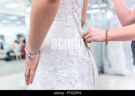 Closeup back of young woman and shop assistant trying on laced corset wedding dress in boutique discount store with lacing ribbon, blonde hair Stock Photo