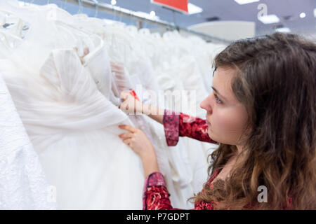 Young woman searching, trying on, choosing, touching wedding dresses in boutique discount store, many white garments hanging on rack hangers row Stock Photo