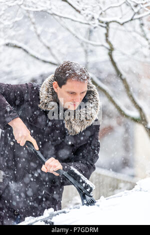 Young man cleaning car windshield from snow, ice with brush and scraper tool during snowfall while snowing snowflakes falling Stock Photo